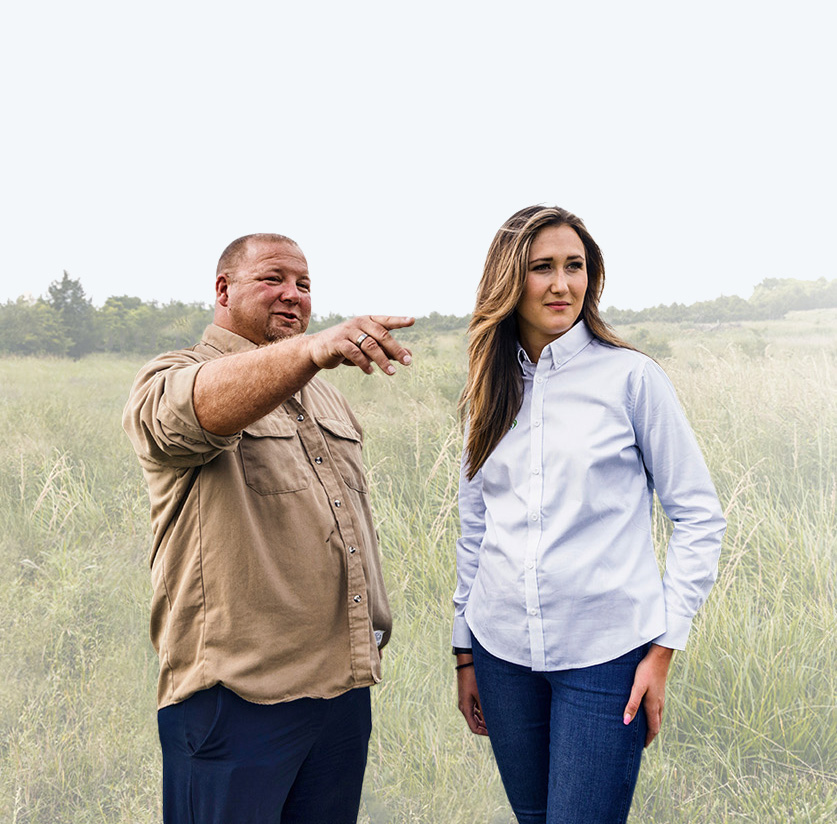 A man points toward the horizon while a woman listens, both standing in a grassy field.